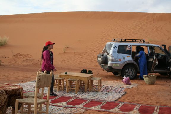 A woman standing near a 4x4 vehicle in a sandy desert setting with a table and chairs.