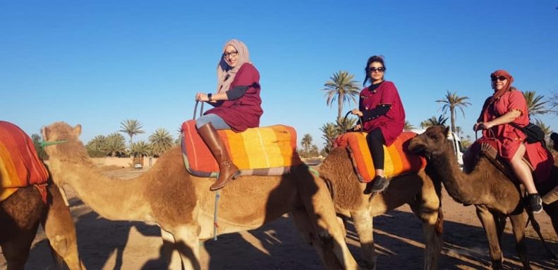 Three people riding camels in the desert, wearing traditional attire and enjoying the sunny day.